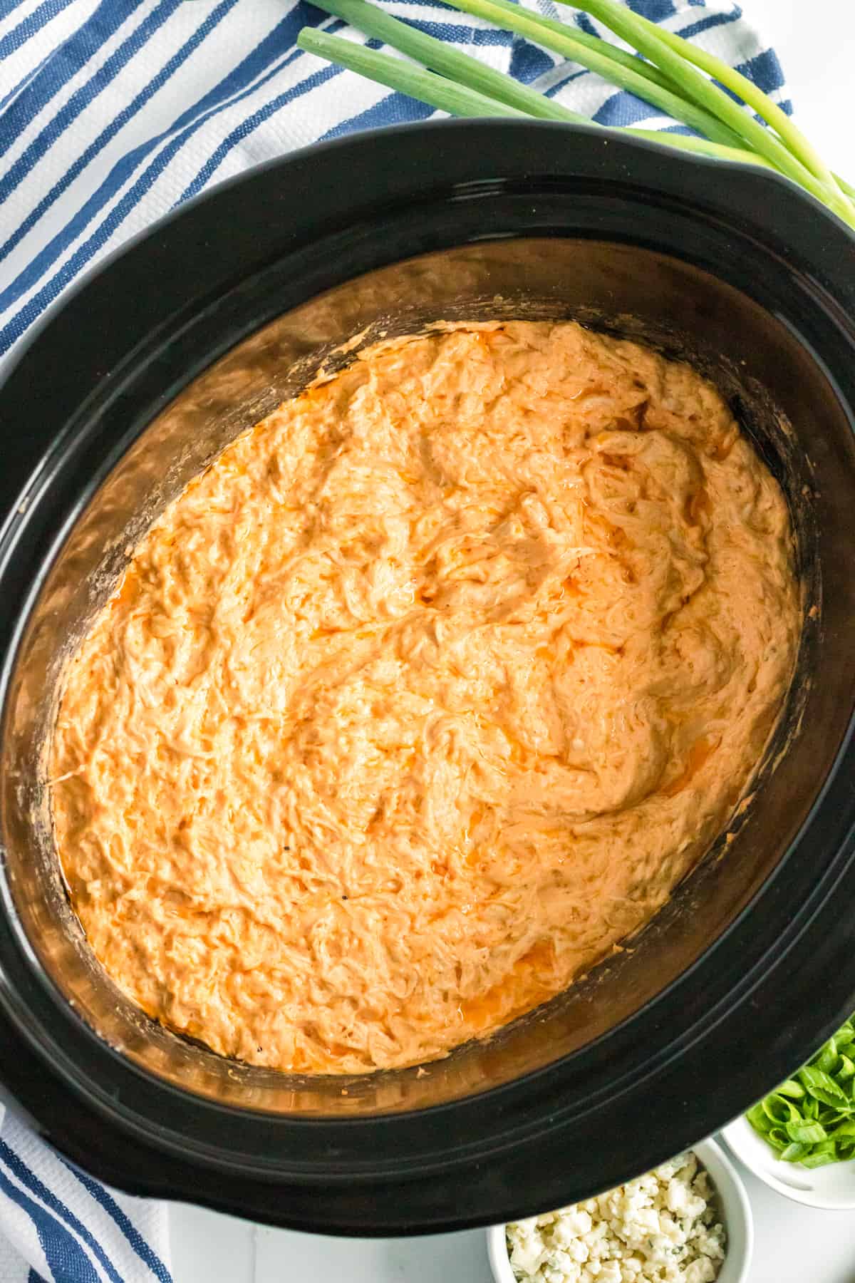 Buffalo chicken dip in the crockpot with green onions and crumbled blue cheese in the background