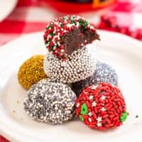 no-bake brownie batter truffles decorated with holiday sprinkles on white plate. On right, brownie truffles stacked on top of one another with a bite taken out of top one to show fudgy inside