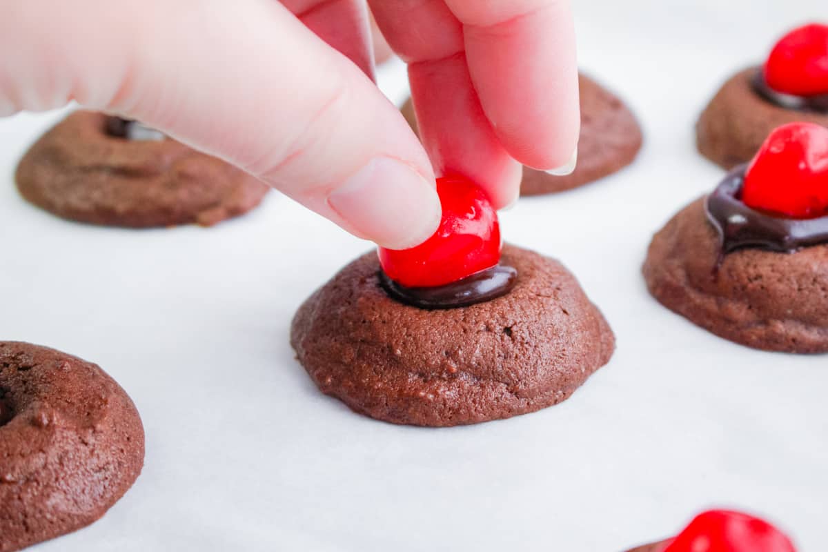 hand placing stemless maraschino cherry on top of chocolate thumbprint cookie