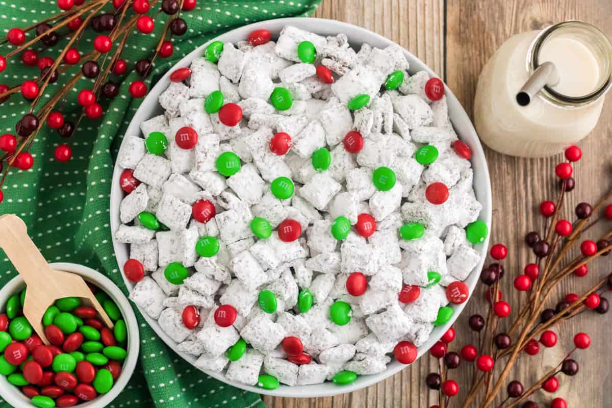 Christmas Puppy Chow with M&Ms in a white bowl. Holiday M&Ms, glass of milk, and holiday decor are surrounding the bowl.