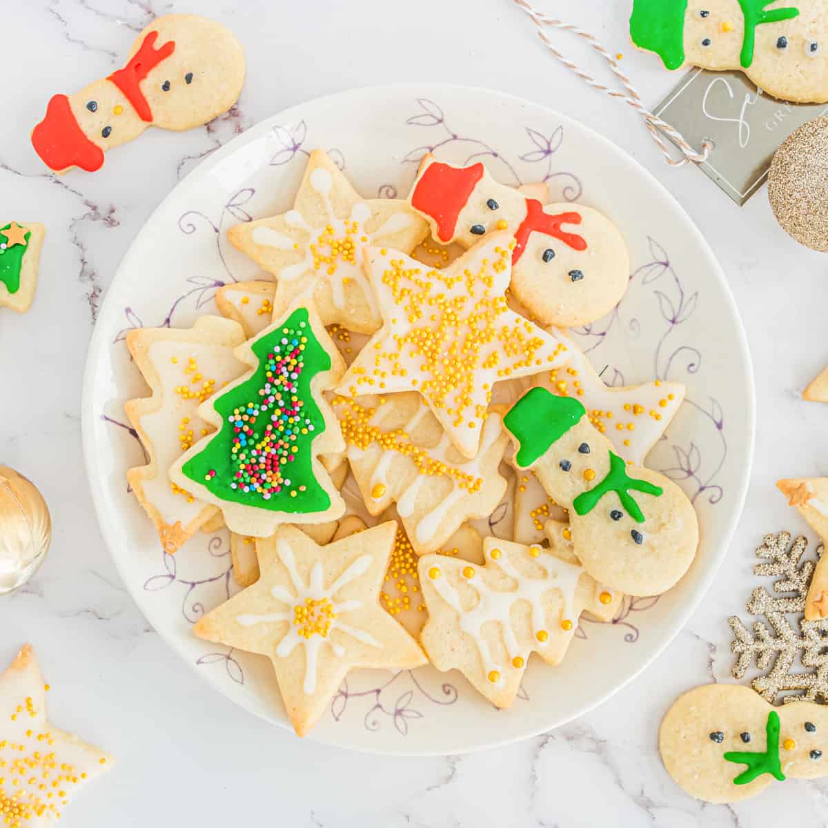 Decorated Christmas sugar cookies with icing and sprinkles on serving plate with additional cookies scattered around table