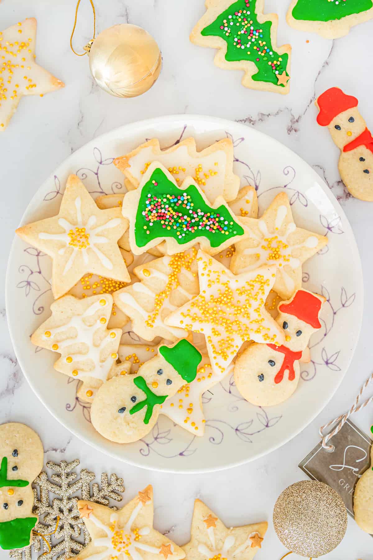 Decorated Christmas sugar cookies with icing and sprinkles on serving plate with additional cookies and Christmas ball ornaments scattered around table