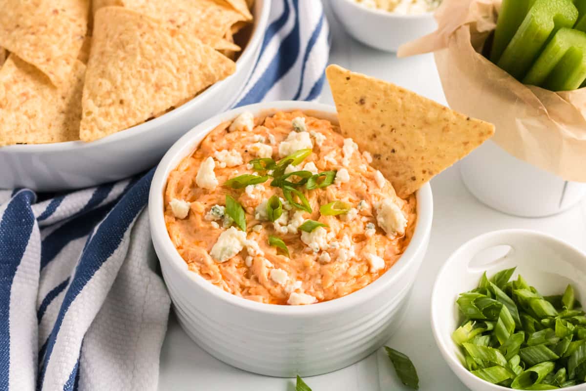 Slow Cooker Buffalo Chicken Dip in white bowl garnished with blue cheese and green onions. A tortilla chip is sticking out of the top.