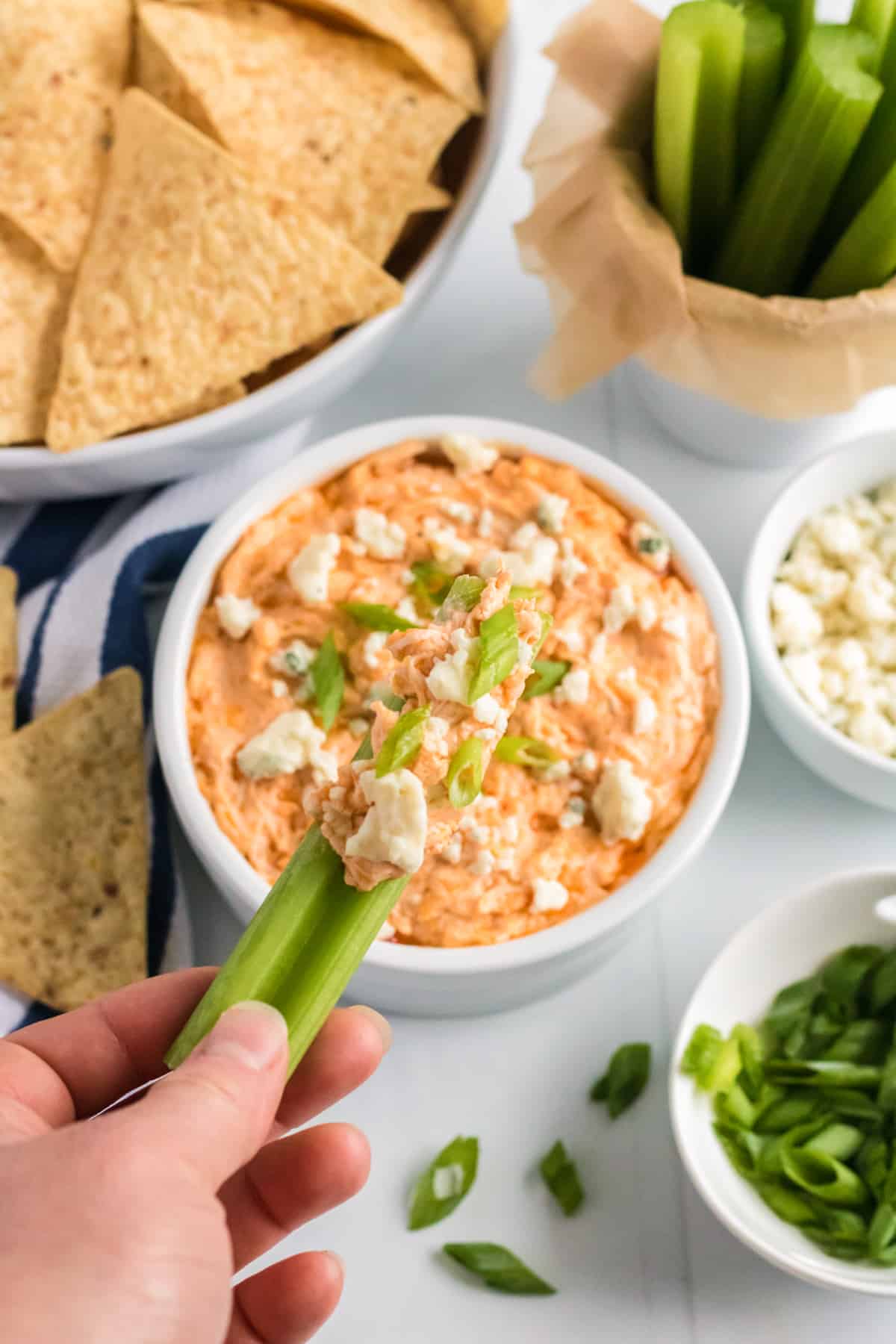 Celery stick being dipped in a bowl of cheesy buffalo chicken dip. Tortilla chips, more celery sticks, blue cheese crumbles, and scallions are in background.