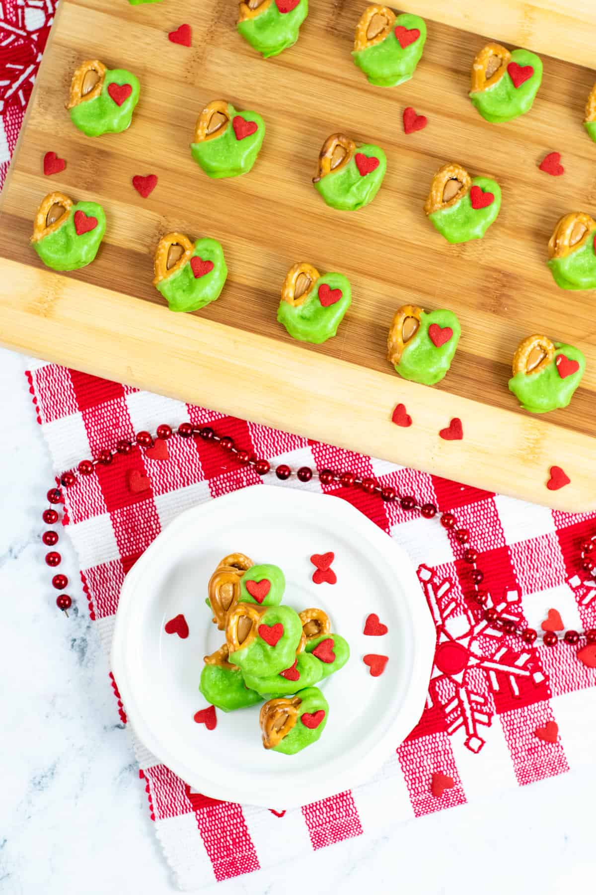 Grinch Pretzel Bites on white plate with red heart sprinkles sprinkled around them and a tray of neatly arranged pretzel bites in background