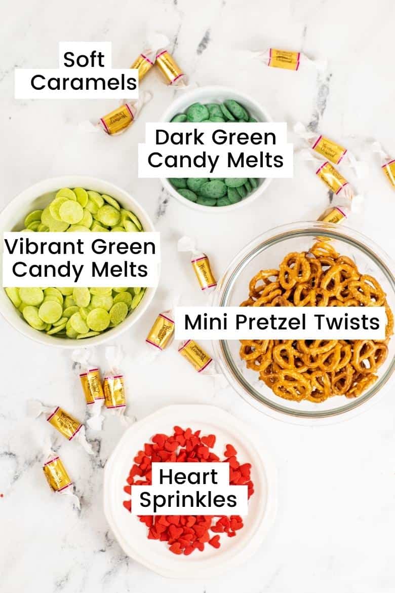 Ingredients in bowls: vibrant green candy melts, dark green candy bites, ini pretzel twists, red hart sprinkles, and soft caramels