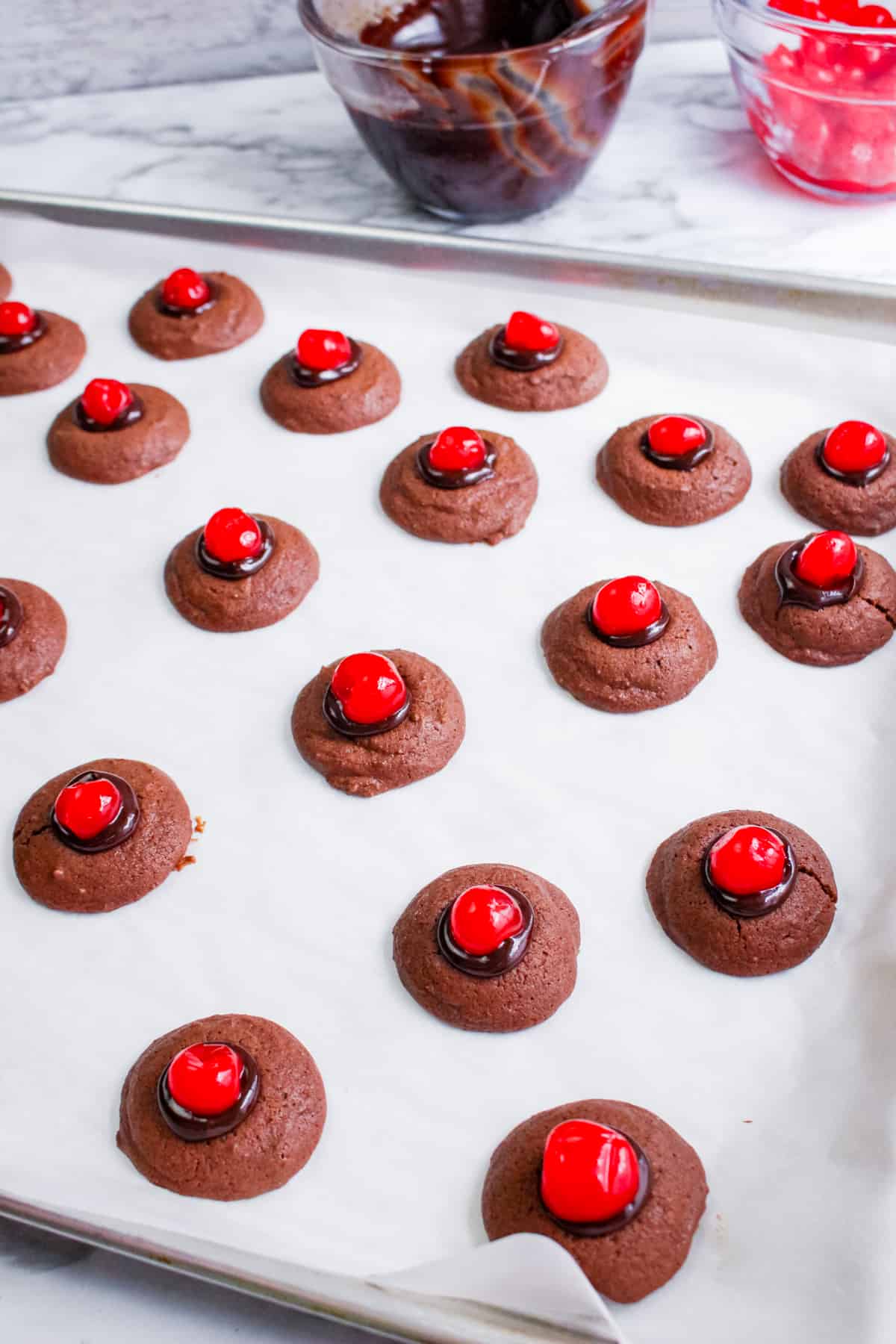 lined baking sheet with small chocolate thumbprint cookies topped with melted chocolate and maraschino cherries