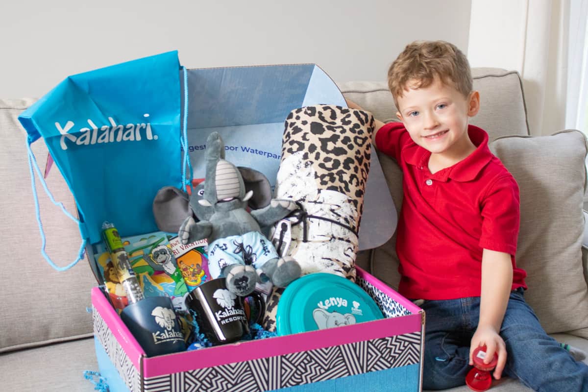 Box of Kalahari Resort merchandise including blanket, frisbee, mugs, and stuffed elephant. Smiling young boy next to the box.