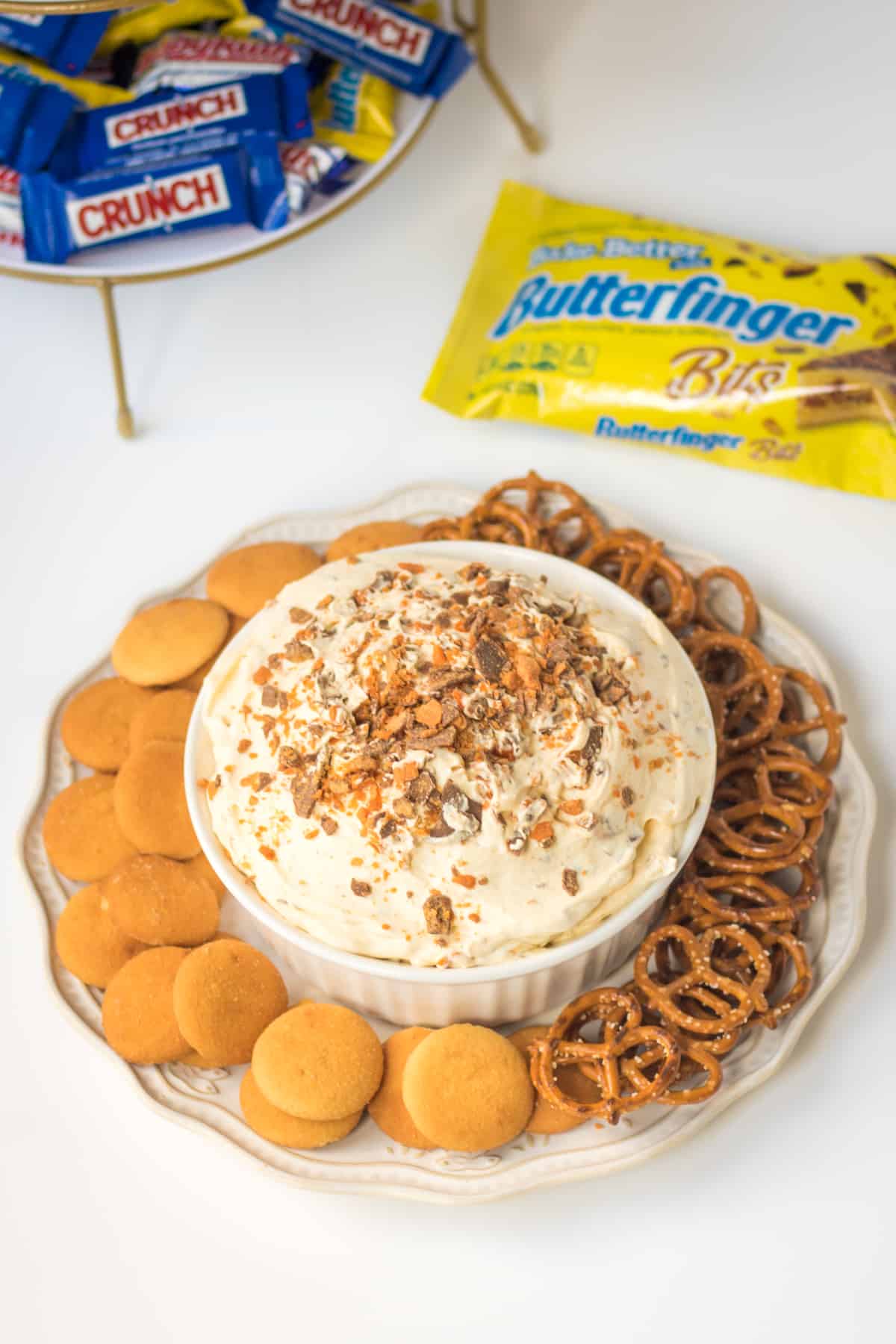 No-bake dessert dip with Butterfinger pieces in it, served with vanilla wafer cookies and pretzel twists for dipping. Bag of Butterfinger Baking bits and fun size Crunch and Butterfinger bars in background