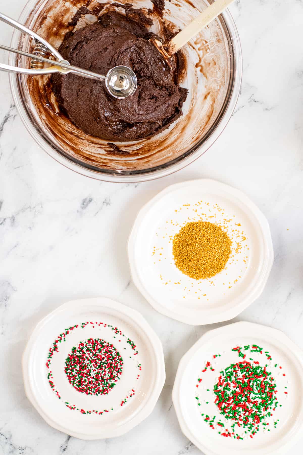 bowl of no-bake brownie batter with cookie scoop in it and three shallow bowls of different color sprinkles.