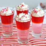 Four Valentine's Day Jello Shots with layers of red and pink gelatin and topped with whipped cream and Valentine's Day Sprinkles