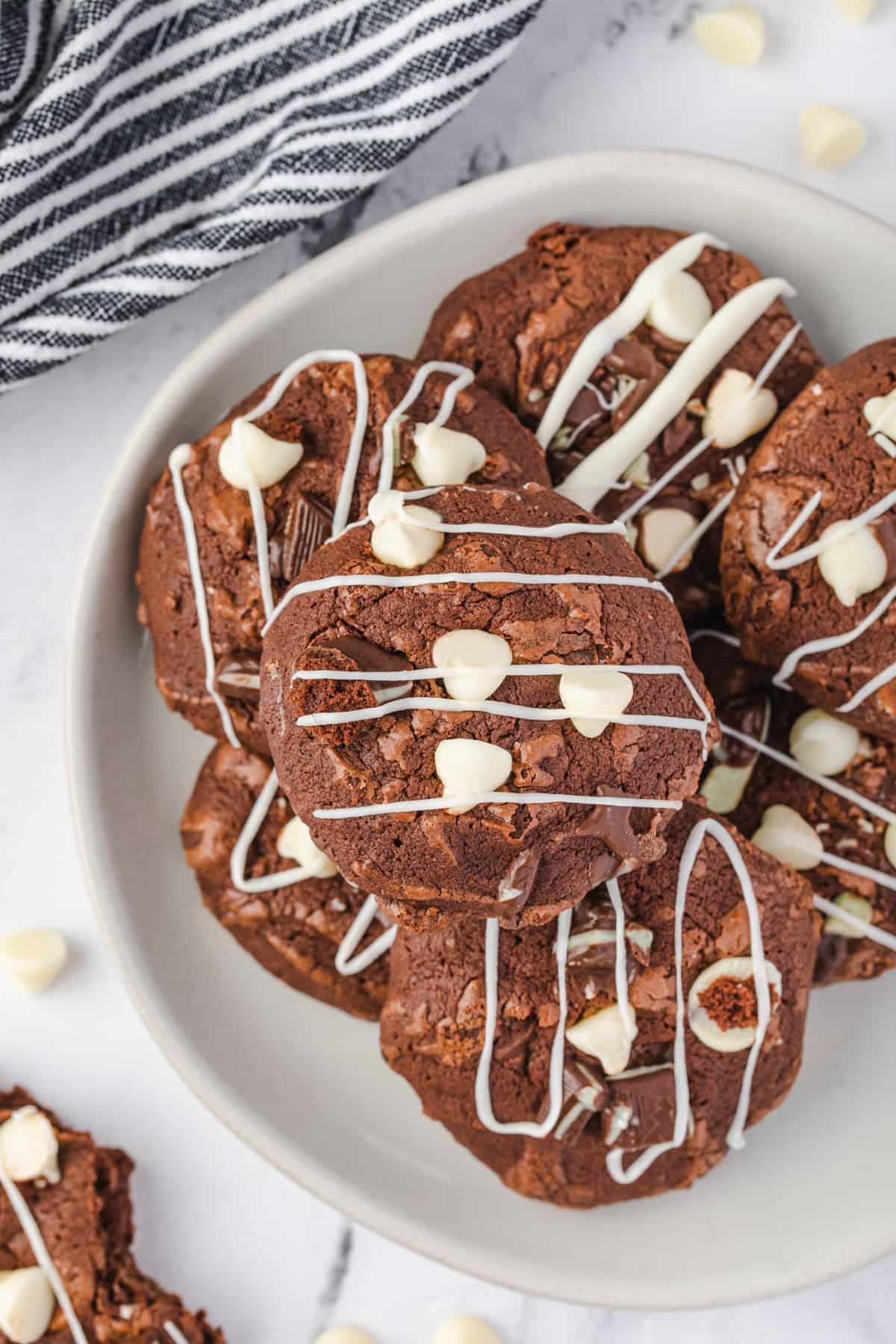 Brownie Cookies with White Chocolate Chips and white chocolate drizzle piled on a serving plate.