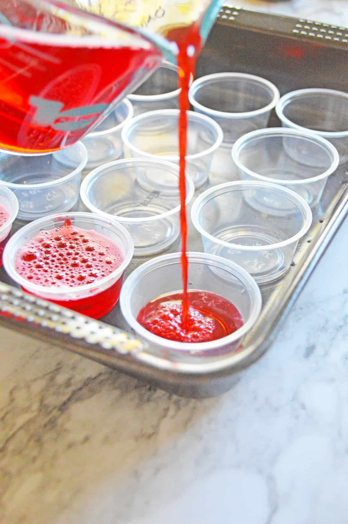 Jello shots being poured into plastic shot cups in a baking pan