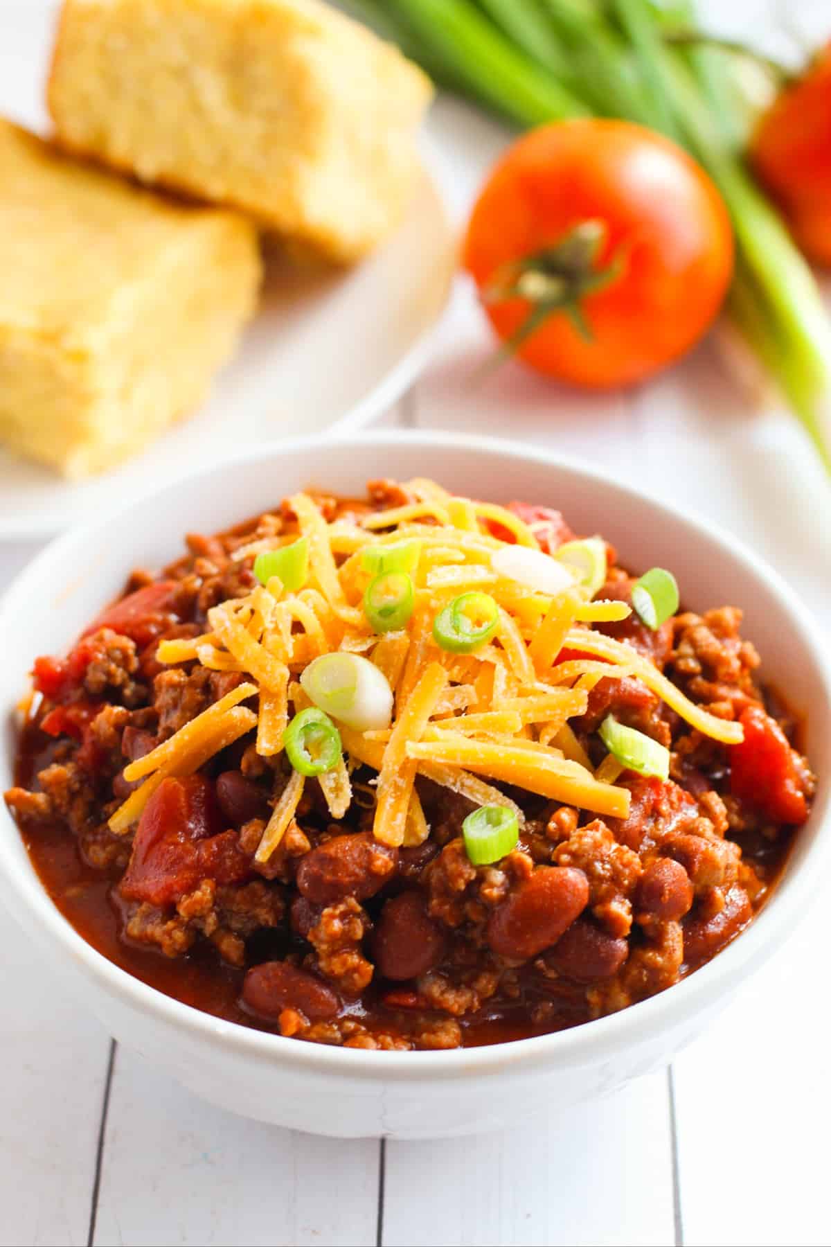 bowl of chili with beans and ground beef topped with shredded cheese and scallions. A tomato, scallions, and rolls are in the background.