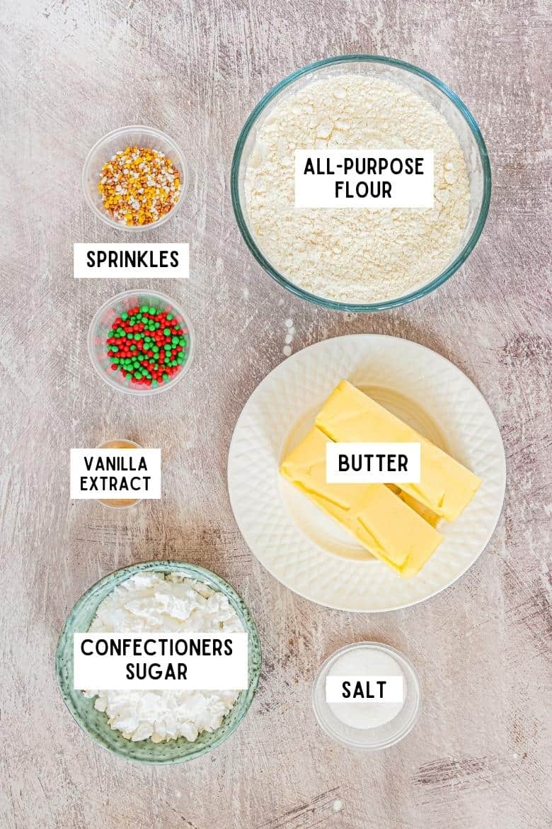 Bowl of all-purpose flour, two sticks of butter, bowl of salt, bowl of confectioners sugar, cup of vanilla extract and to bowls of holiday sprinkles