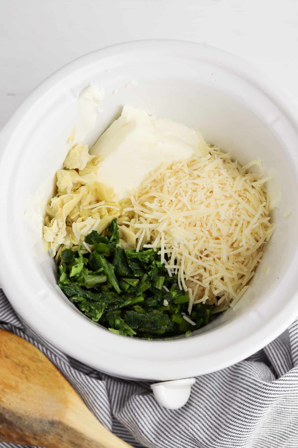 Cream cheese, spinach, artichokes, garlic, and shredded cheese in round white crockpot