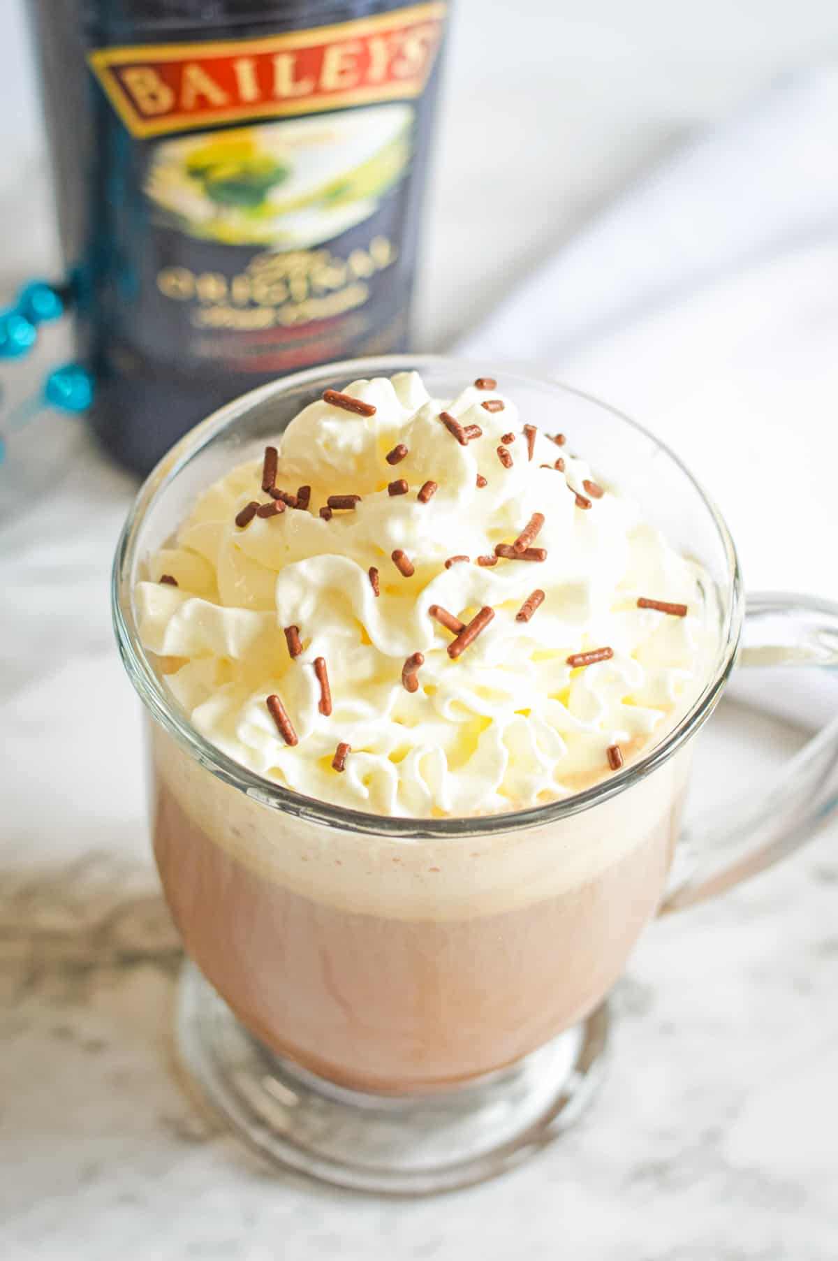 Dirty snowman adult hot chocolate cocktail with whipped cream and chocolate sprinkles in a glass mug with a bottle of Baileys in the background.