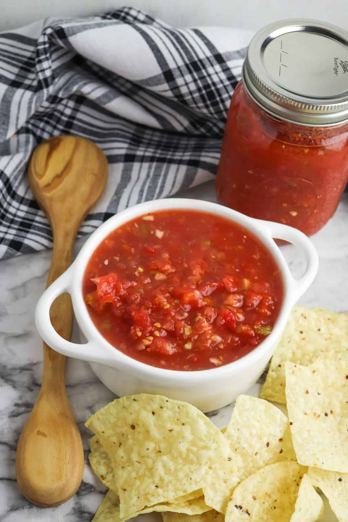 Bowl of homemade salsa with tortilla chips, cloth napkin, wooden spoons, and jar of homemade salsa in background