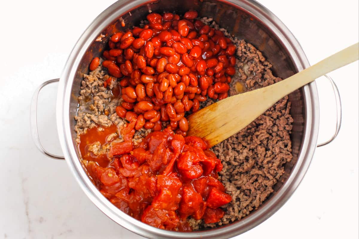large pot with browned ground beef, chili beans, diced tomatoes, and a wooden spoon.