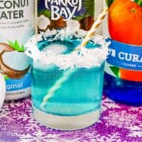 Bright blue jack frost cocktail in rocks glass with coconut flake rim and bottles of coconut water, coconut rum, and blue curaco in background