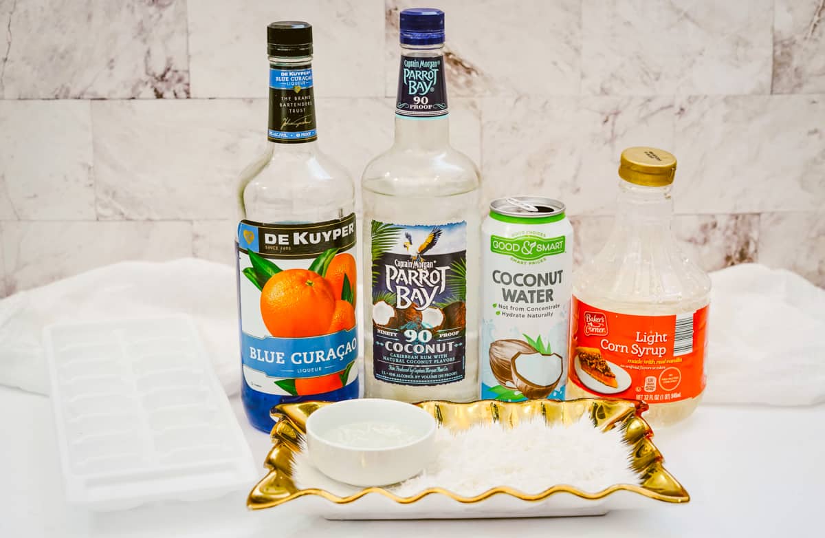tray of ice cubes, bottle of blue Curaçao, bottle of captain morgan parrot bay, can of coconut water, bottle of light corn syrup, shallow platter with coconut flakes