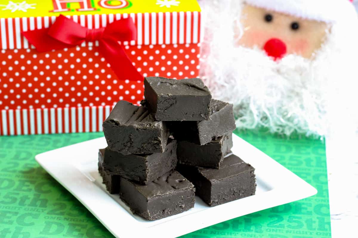 Square pieces of black fudge piled on a white plate with santa and other christmas decorations in the background.