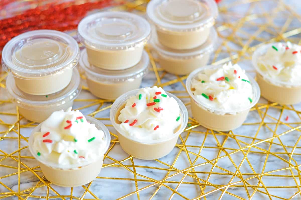christmas cookie jello shots with whipped cream and sprinkles lined up in a row in front of additional shots in cups with covers on them