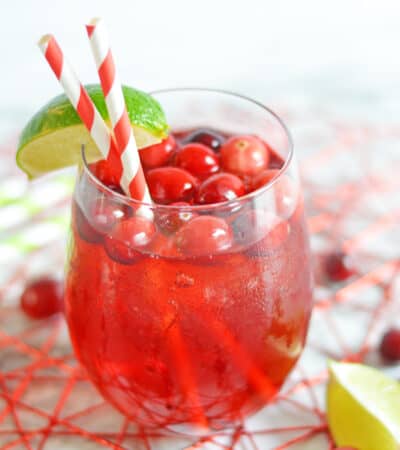 Vodka Cranberry Cocktail in short glass with ice, fresh cranberries, paper straws, and lime wedge on rim.