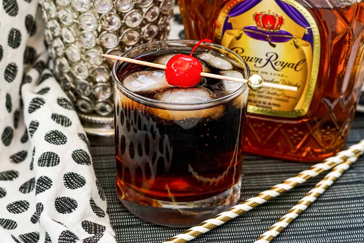 Whiskey Cherry Coke Cocktail in glass with ice cubes and maraschino cherry on skewer, with crown royal bottle behind the glass