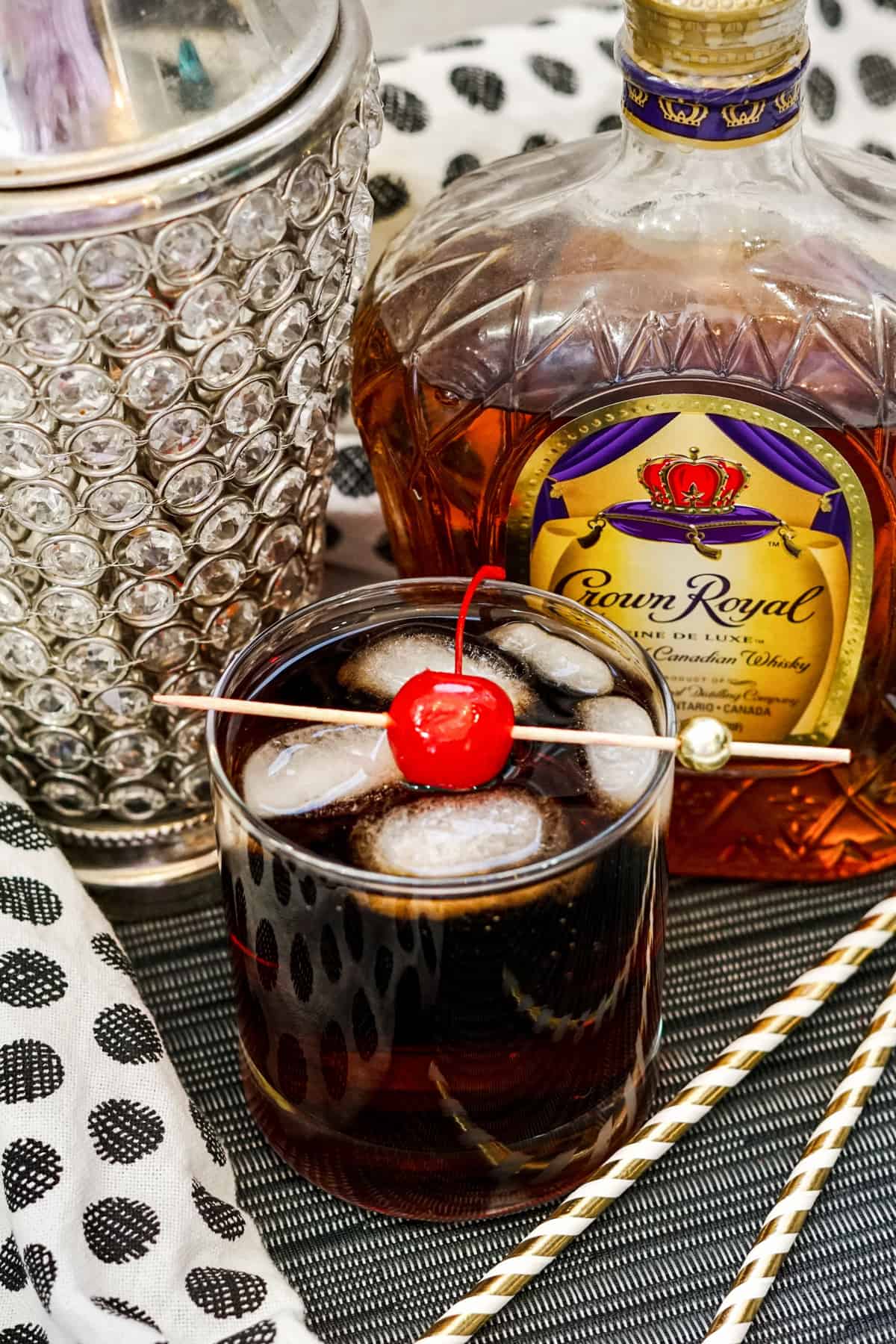 Whiskey Cherry Coke Cocktail in glass with ice and maraschino cherry on skewer and bottle of Crown Royal in background