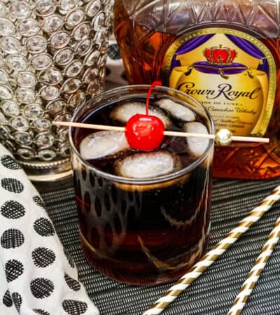 Whiskey Cherry Coke Cocktail in glass with ice cubes and maraschino cherry on skewer. Bottle of crown royal in background.