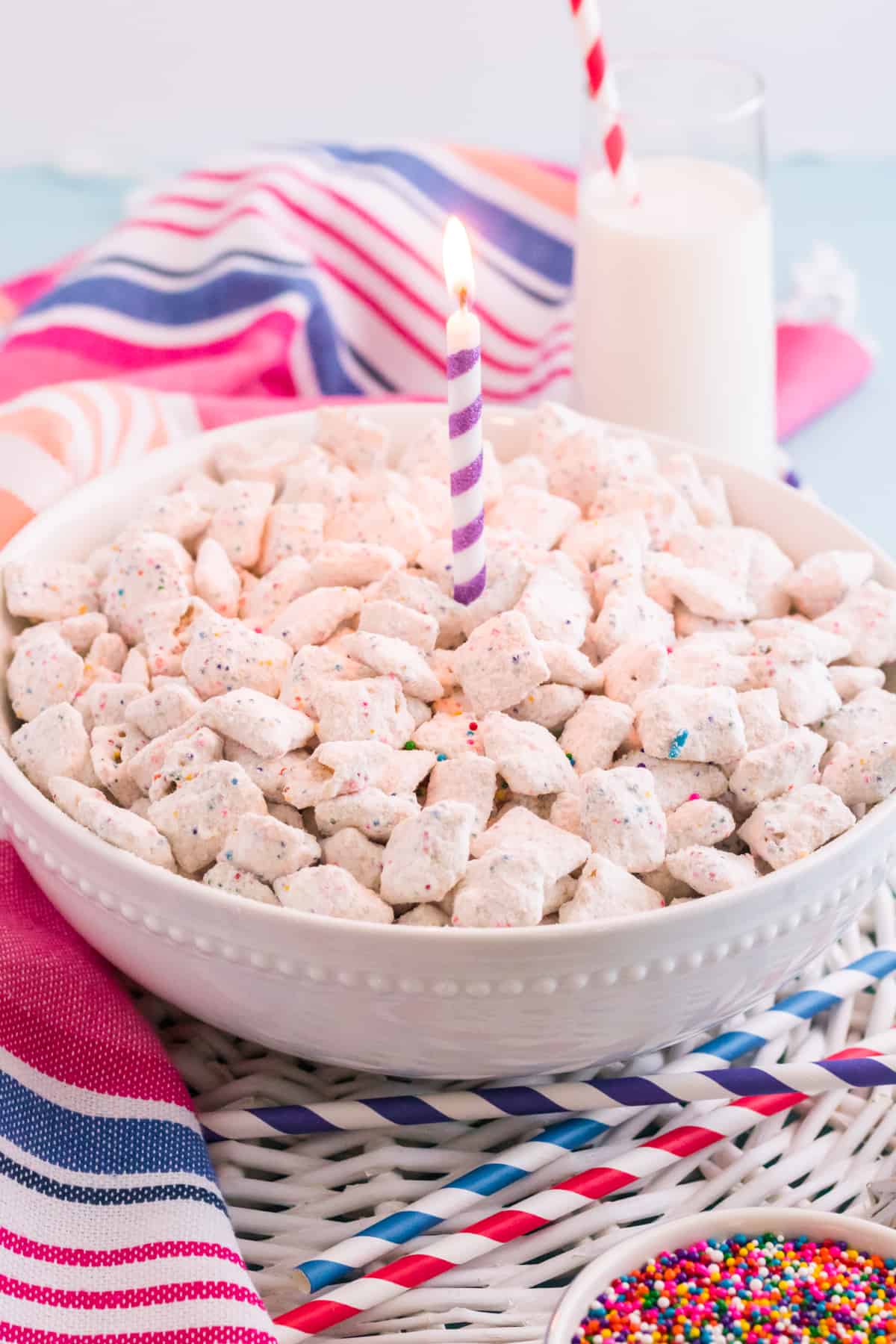 Large white bowl of funfetti puppy chow with purple birthday candle in the center and a glass of milk, colorful paper straws, sprinkles, and a colorful linen in background.