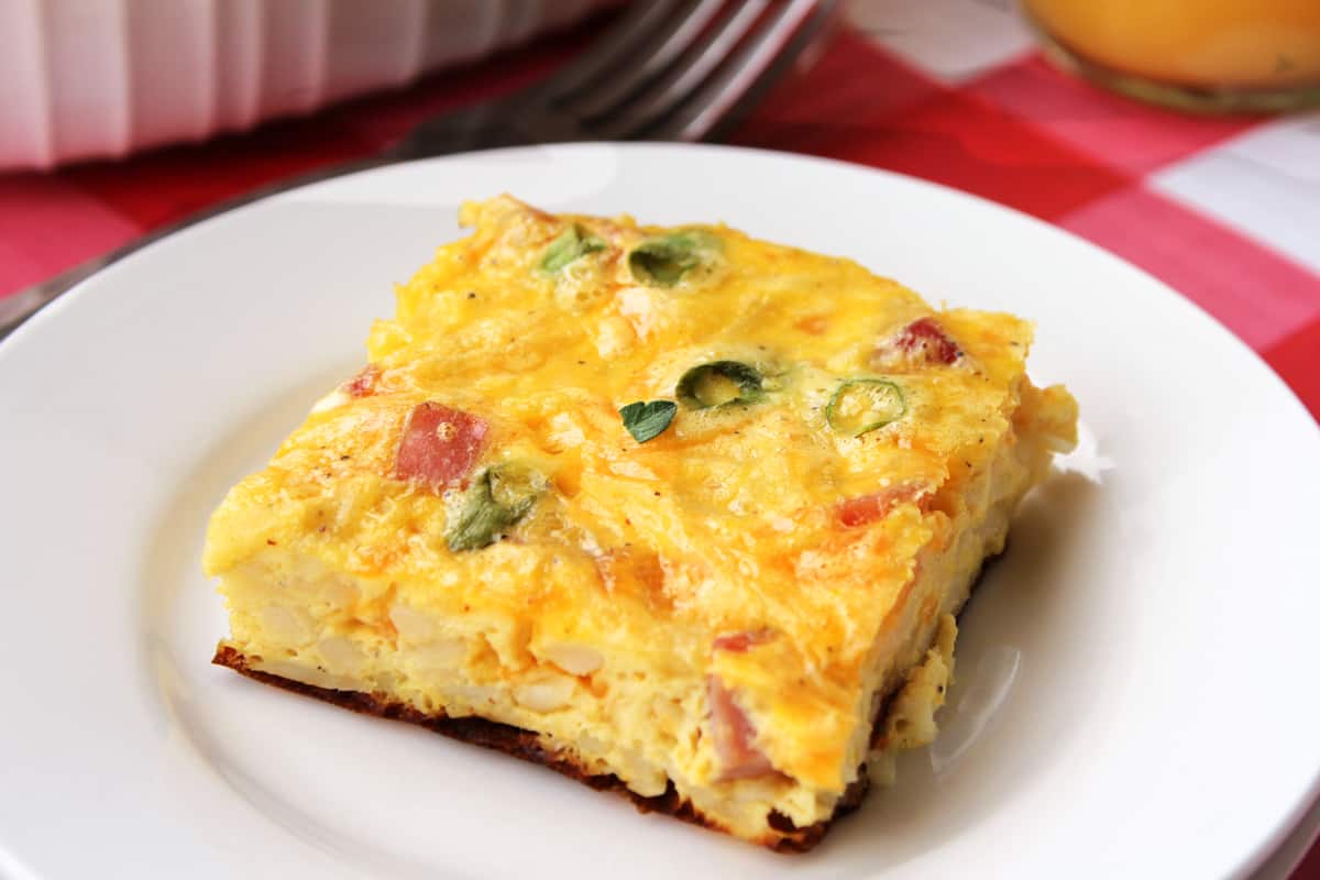 Square slice of breakfast casserole with hash browns, ham, eggs, and green onion served on a white plate with a fork next to it