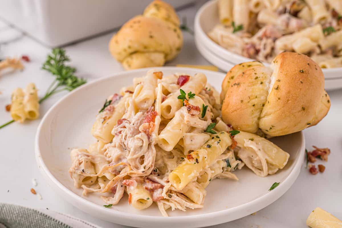 Serving of cheesy chicken alfredo pasta bake on a white plate with a roll and an additional plate and roll in the background