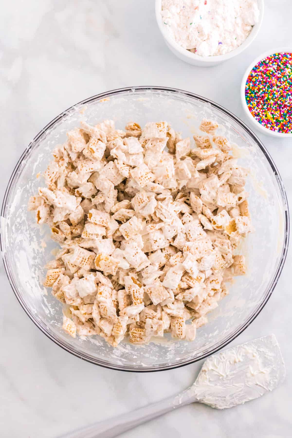 White candy melt coated chex cereal in large glass bowl with rainbow sprinkles, spatula, and funfetti cake mix beside it