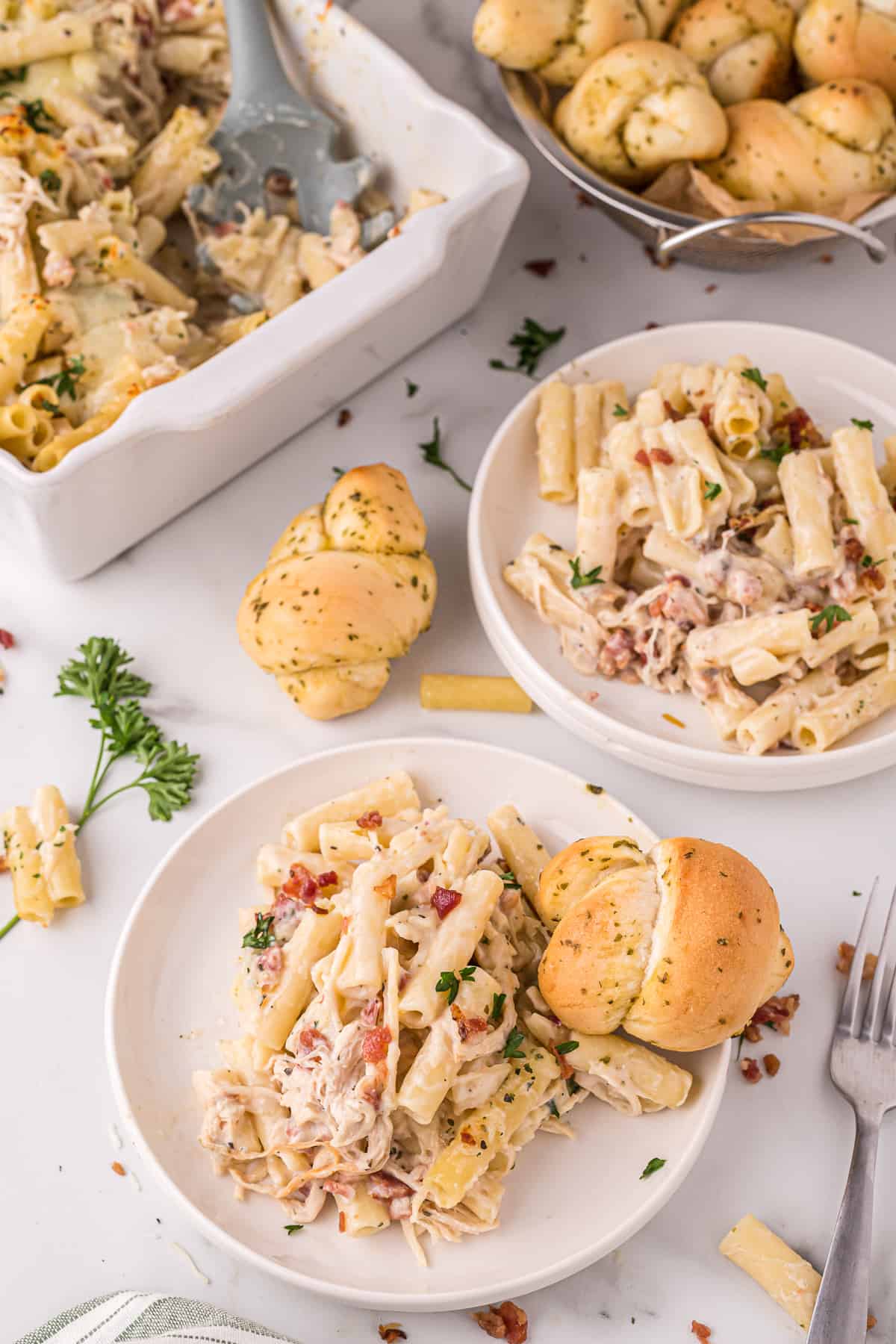 Serving of chicken bacon ziti pasta bake on a white plate with a garlic knot and an additional plated portion, more rolls, and casserole dish in the background 