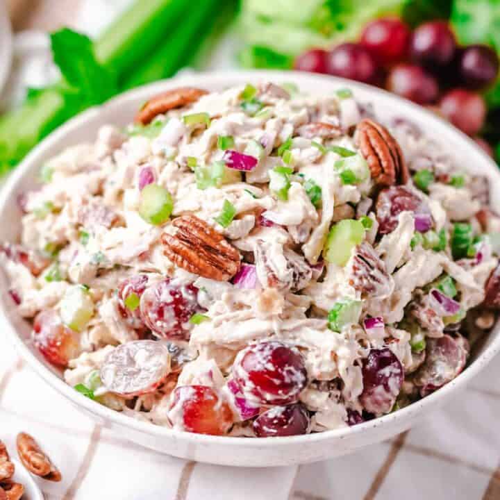 Chicken grape and pecan salad in a white bowl with fresh red grapes, celery, and lettuce behind it