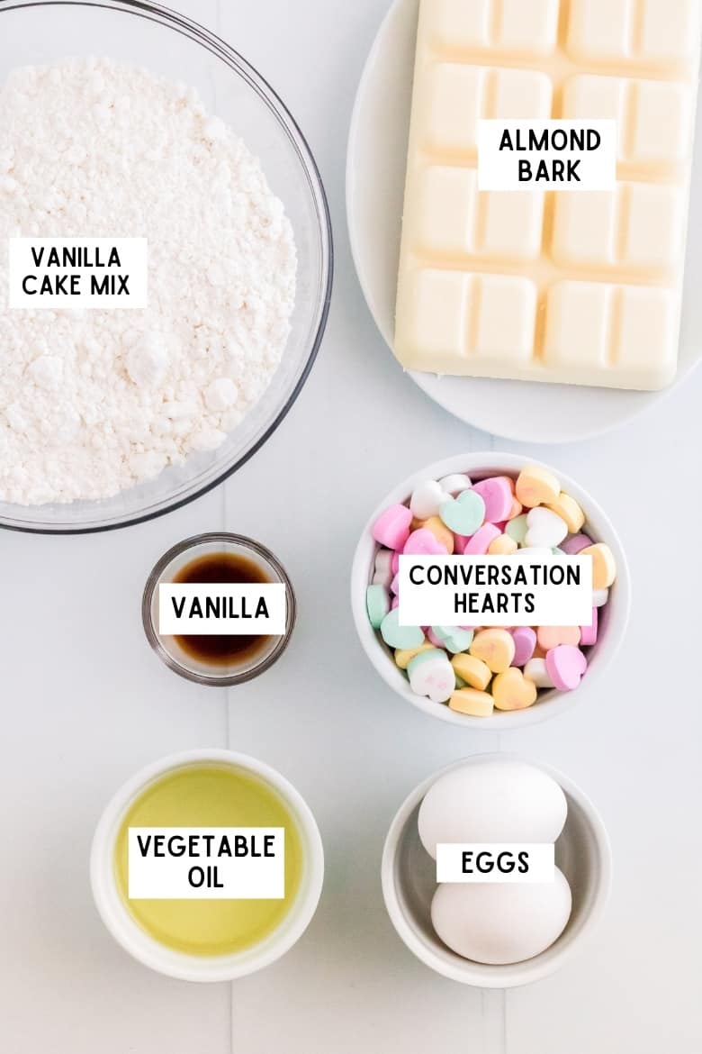 Bowl of vanilla cake mix, blocks of almond bark, bowl of conversation heart candies, bowl of vanilla extract, 2 large eggs, bowl of vegetable oil