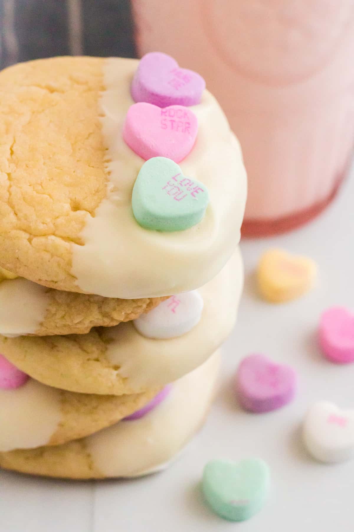 Five Valentines cake mix cookies with candy hearts stacked on top of one another with a glass of milk and more conversation heart candies around them.