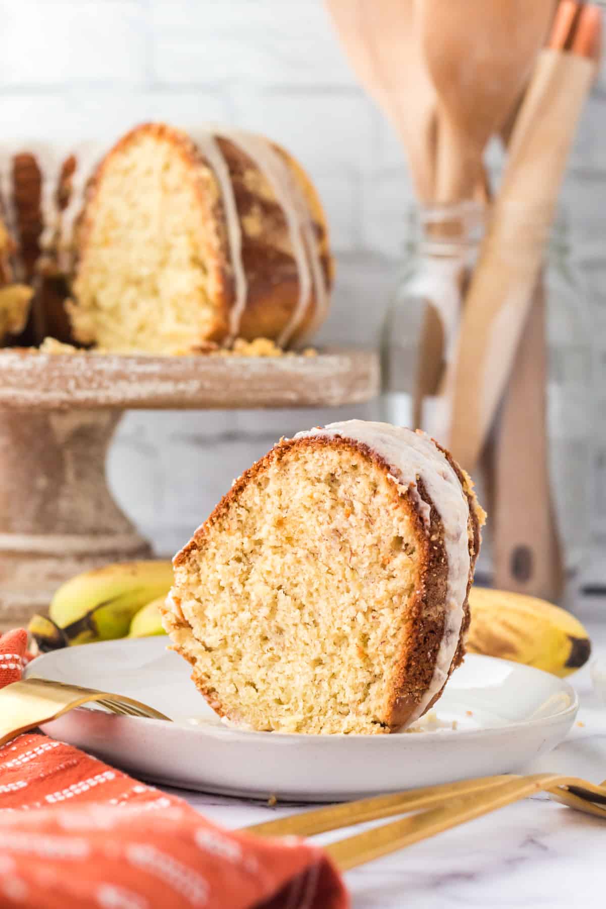 Slice of glazed banana cake on a plate with rest of banana bundt cake on a cake stand in the background.