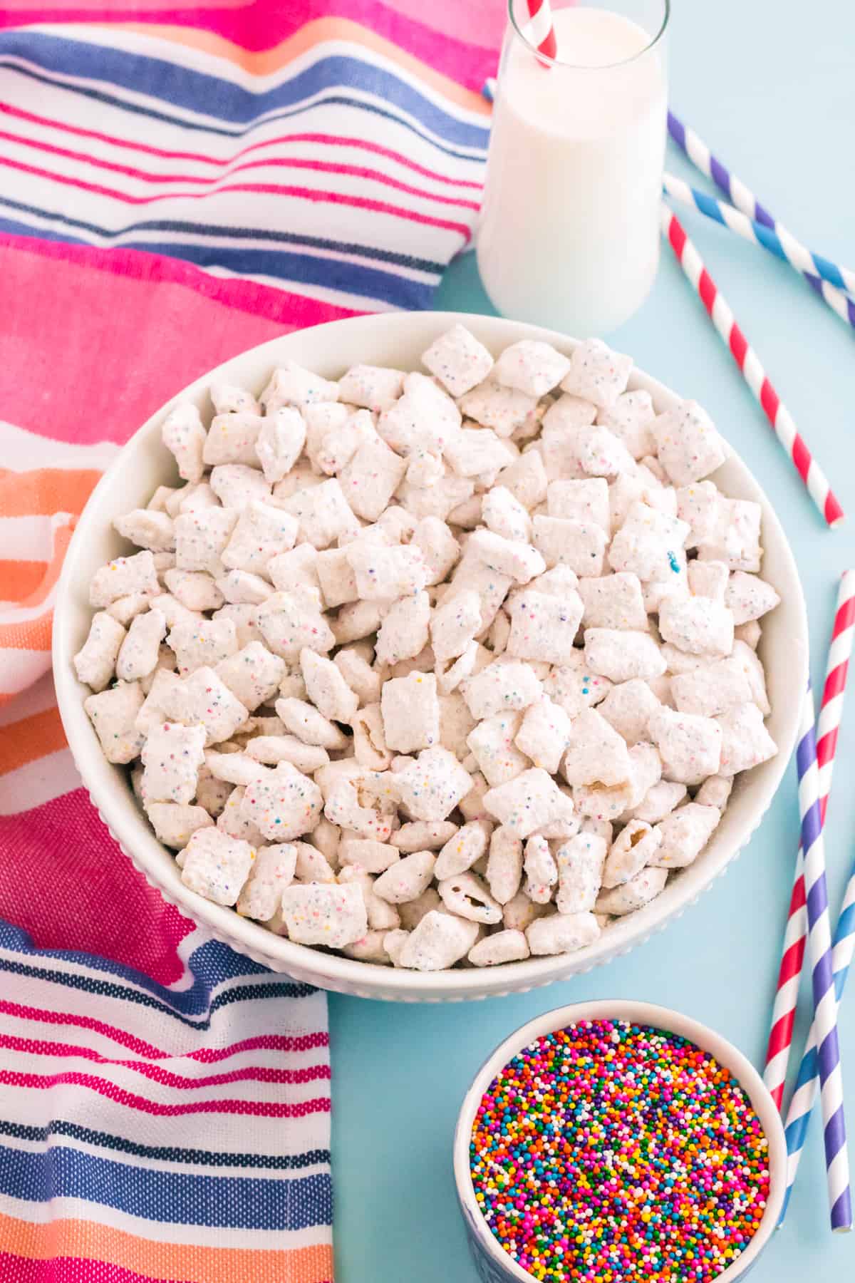 Birthday cake puppy chow in large white bowl with glass of milk, rainbow sprinkles, and colorful linen and straws surrounding it.
