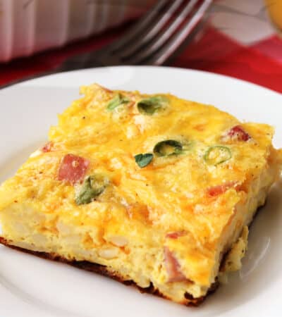 Square slice of breakfast casserole with hash browns, ham, eggs, and green onion served on a white plate with a fork next to it