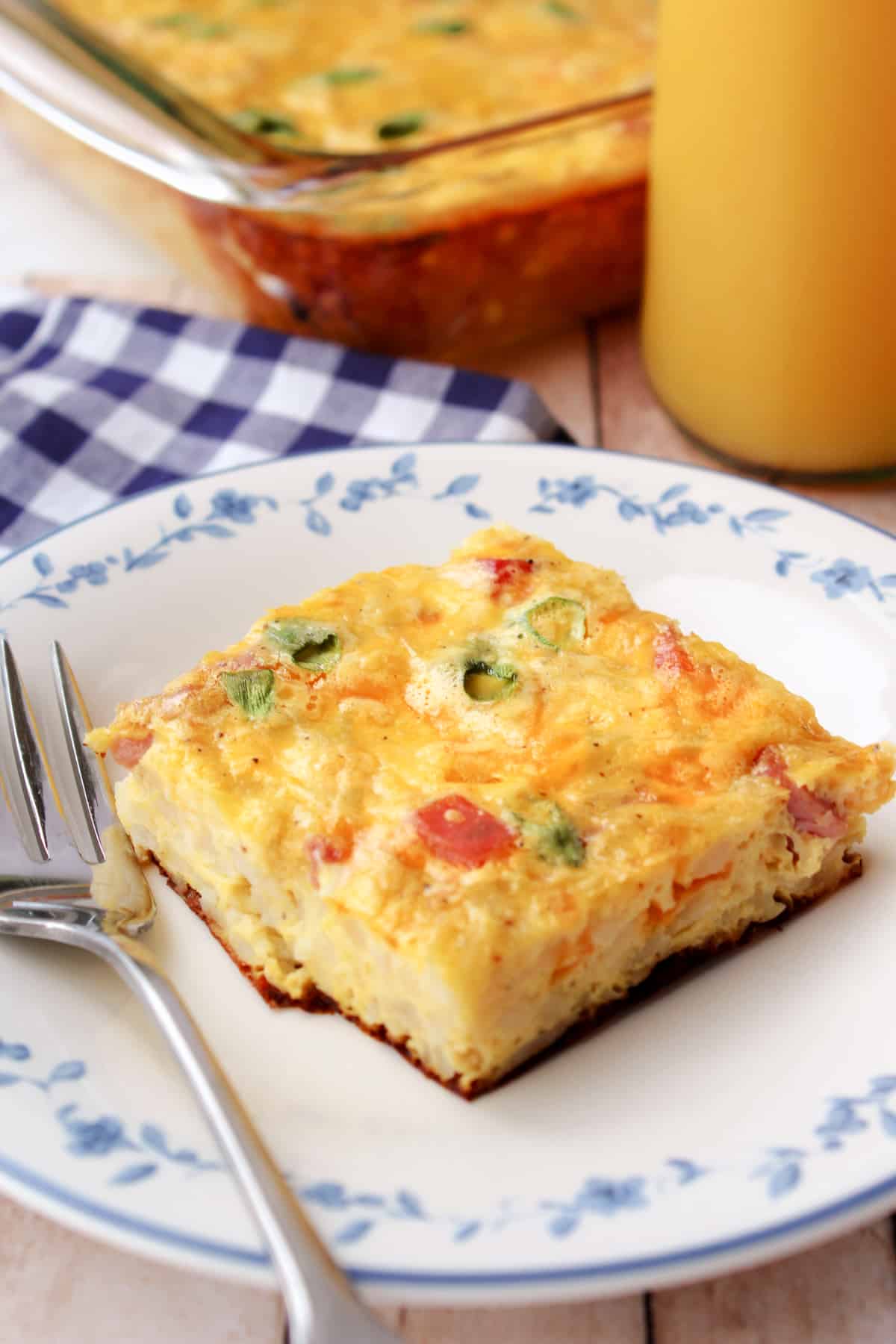 Farmer's Casserole with hash browns and diced ham served on a white and blue plate with a fork, glass of orange juice, and the remaining casserole in the background