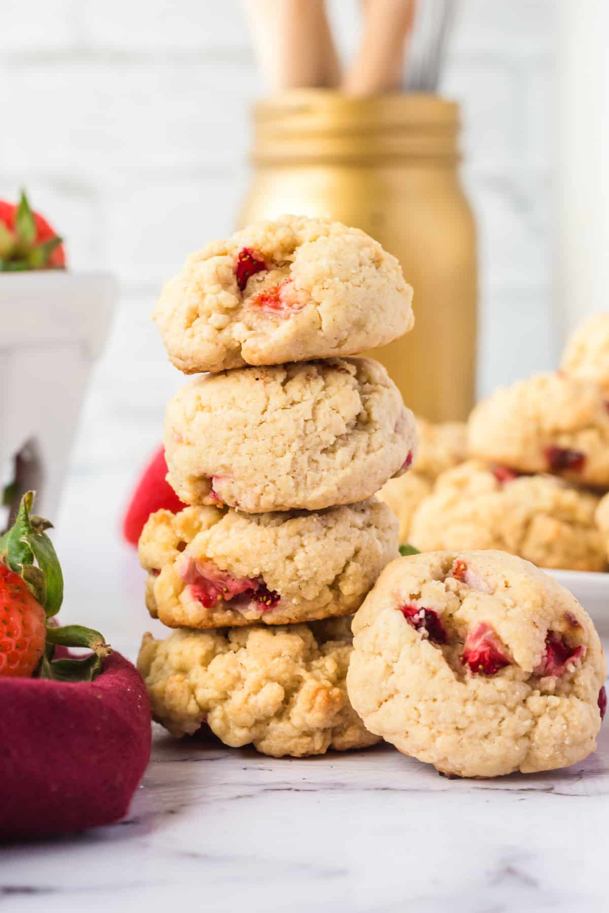 4 fluffy strawberry cookies stacked on top of one another with additional cookies and fresh strawberries surrounding them 