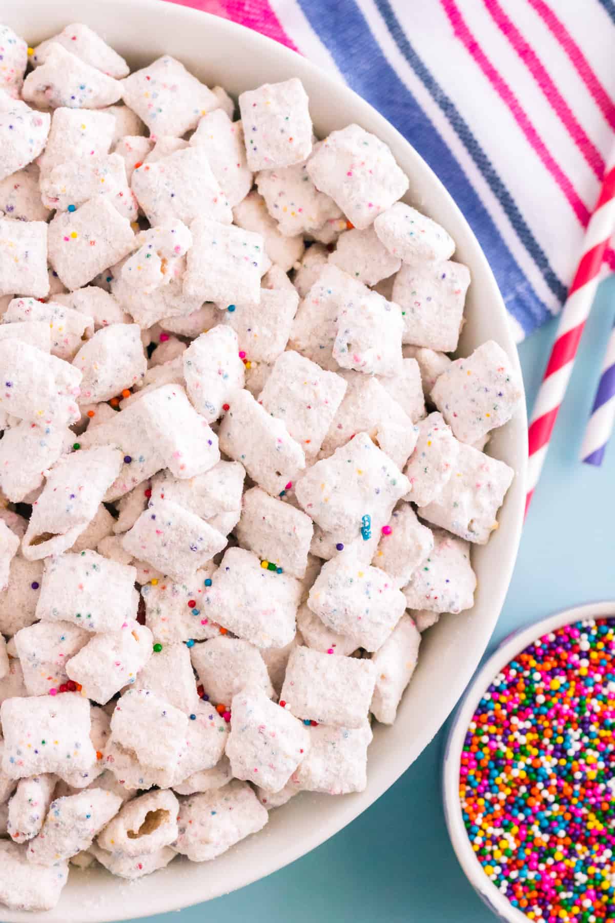 Top-down photo of funfetti chex mix muddy buddies in large white bowl with rainbow sprinkles in a smaller bowl beside them