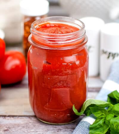 homemade pizza sauce in glass mason jar with tomatoes, salt and pepper, and basil in the background