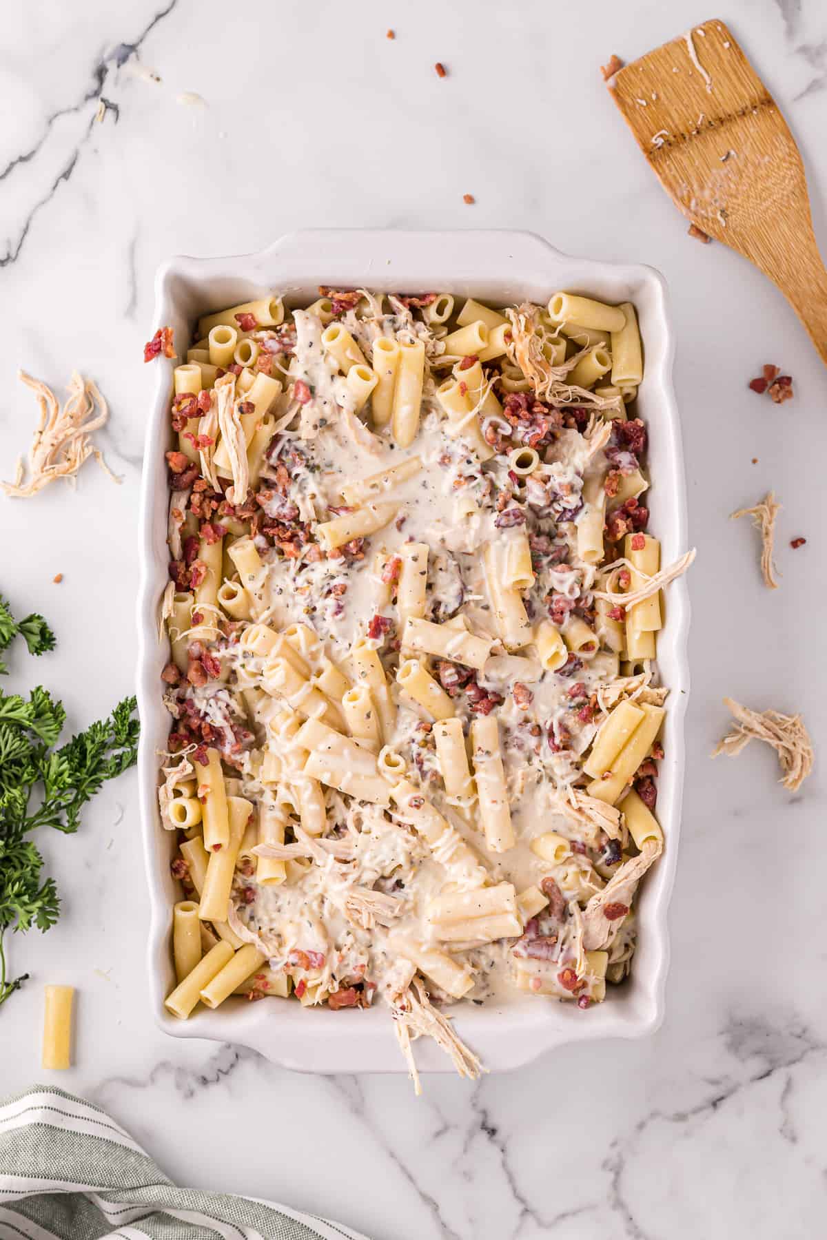 Casserole dish with ziti, crumbled bacon, shredded cooked chicken, and alfredo sauce poured on the top