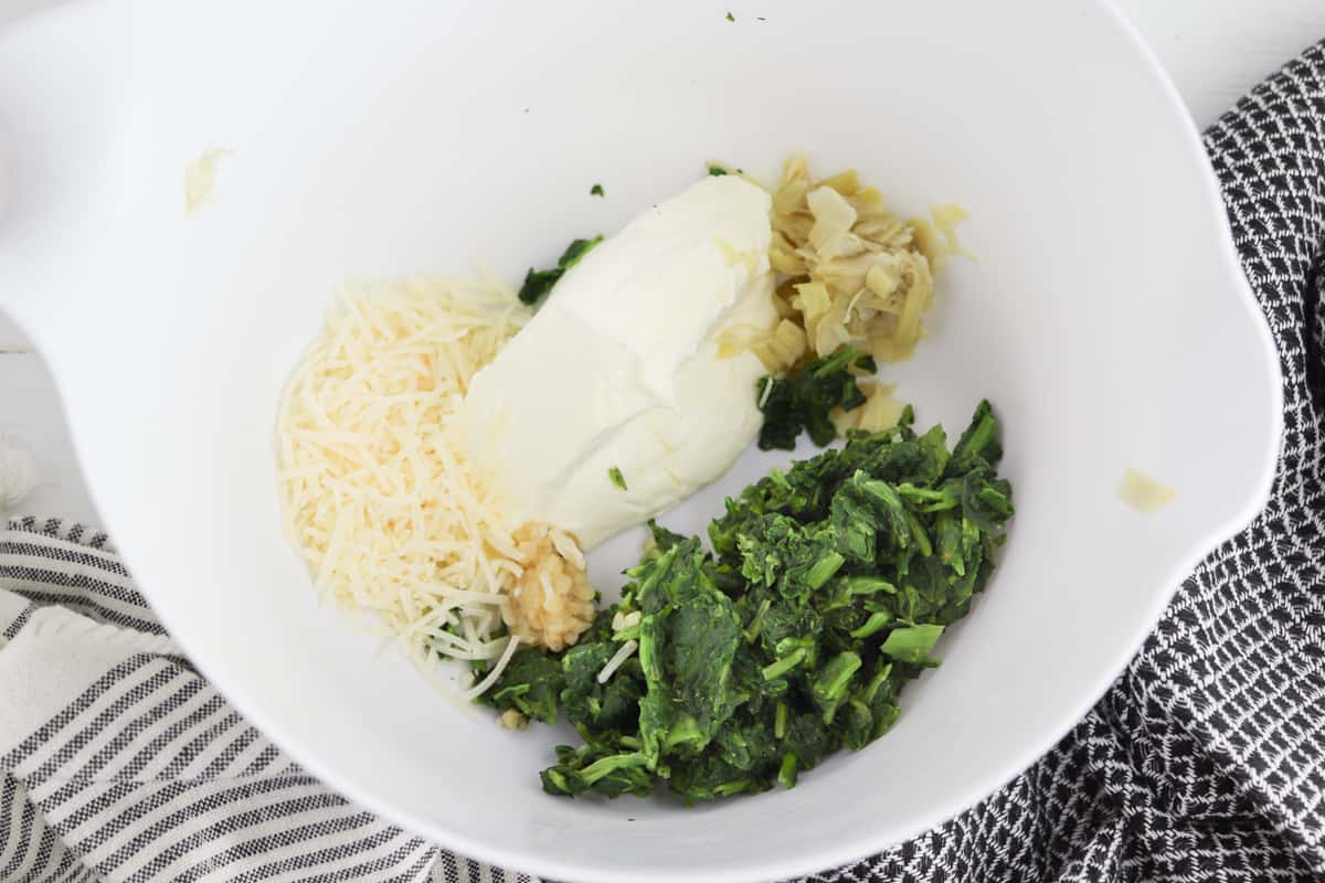 white mixing bowl with chopped spinach, cream cheese, Parmesan cheese, chopped artichoke hearts, minced garlic, and