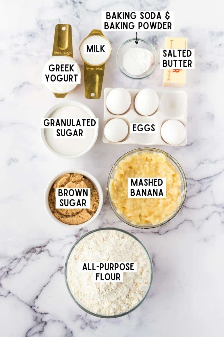 Ingredients on countertop: Greek yogurt, milk, baking powder, baking soda, salted butter, granulated sugar, 4 eggs, brown sugar, mashed banana, and all purpose flour.