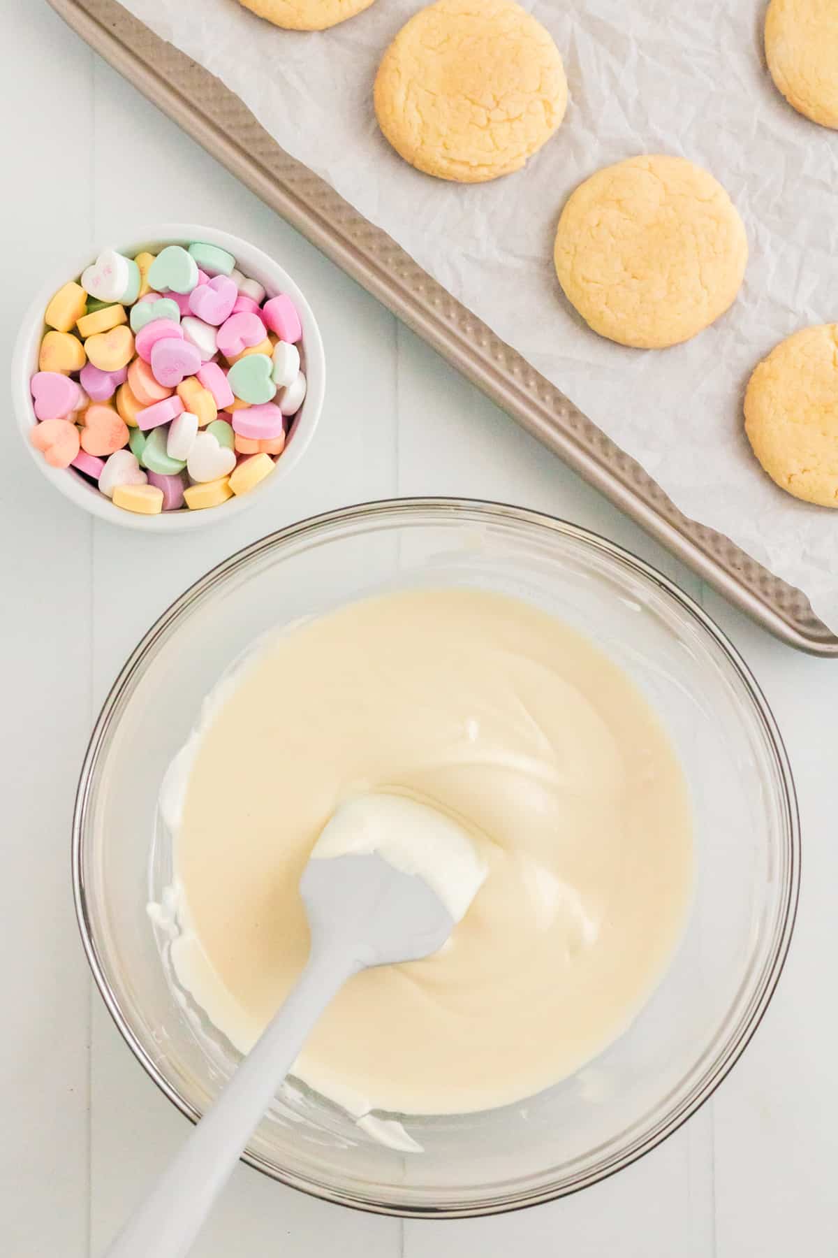 Melted almond bark in glass bowl with candy hearts and vanilla cookies on lined baking sheet in background